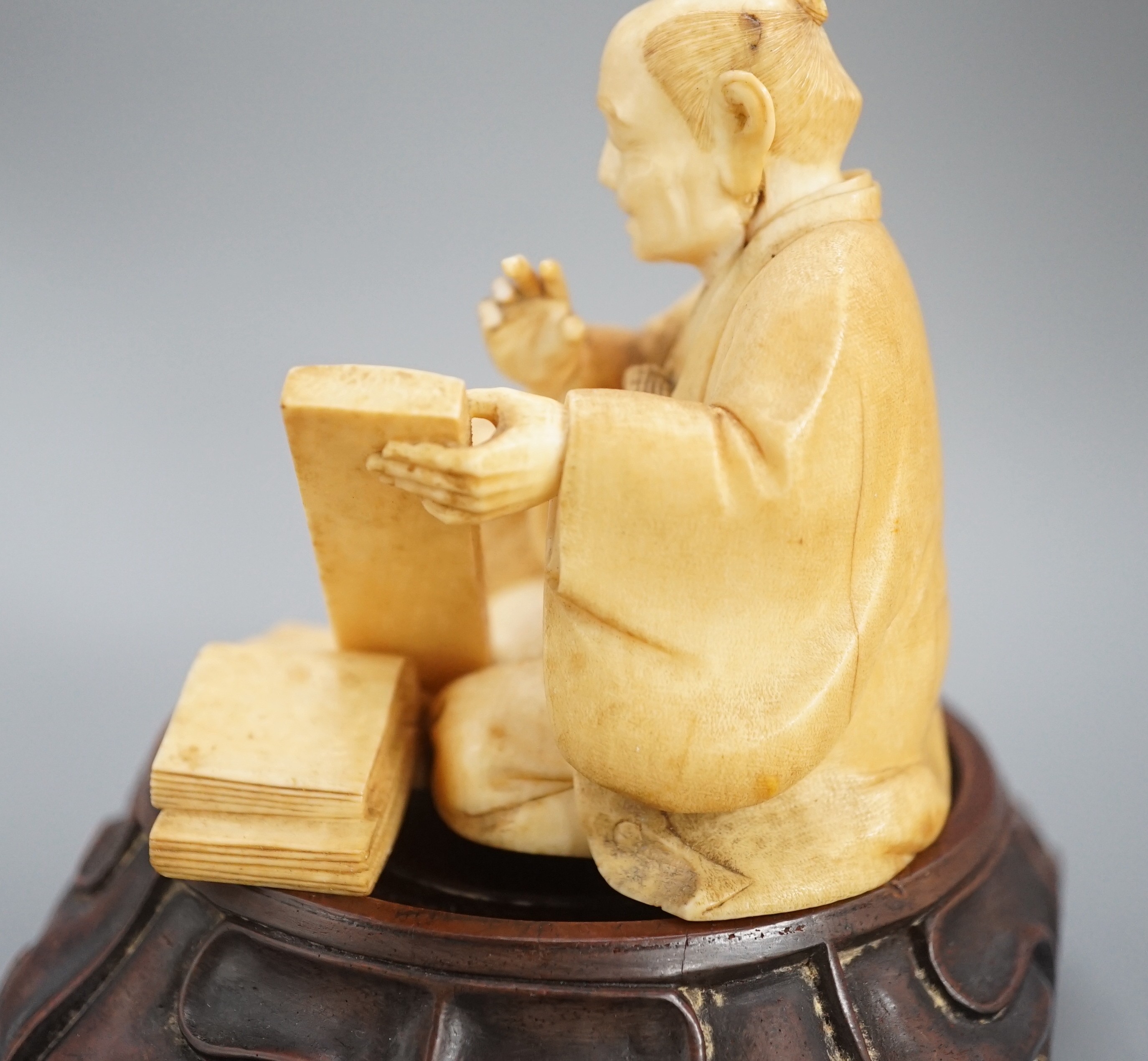 A Japanese ivory okimono of a street vendor kneeling and holding an abacus, Meiji period, 8cm, associated wood stand